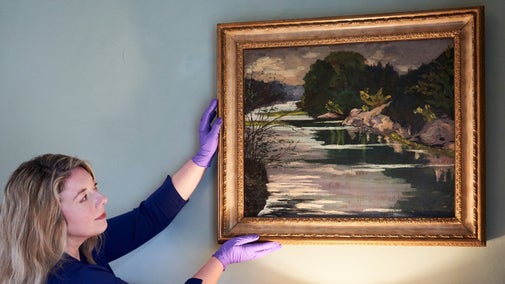 A curator wearing purple gloves, holds the paining 'Quiet Waters' by Churchill against a plain wall. The painting is framed in gold.